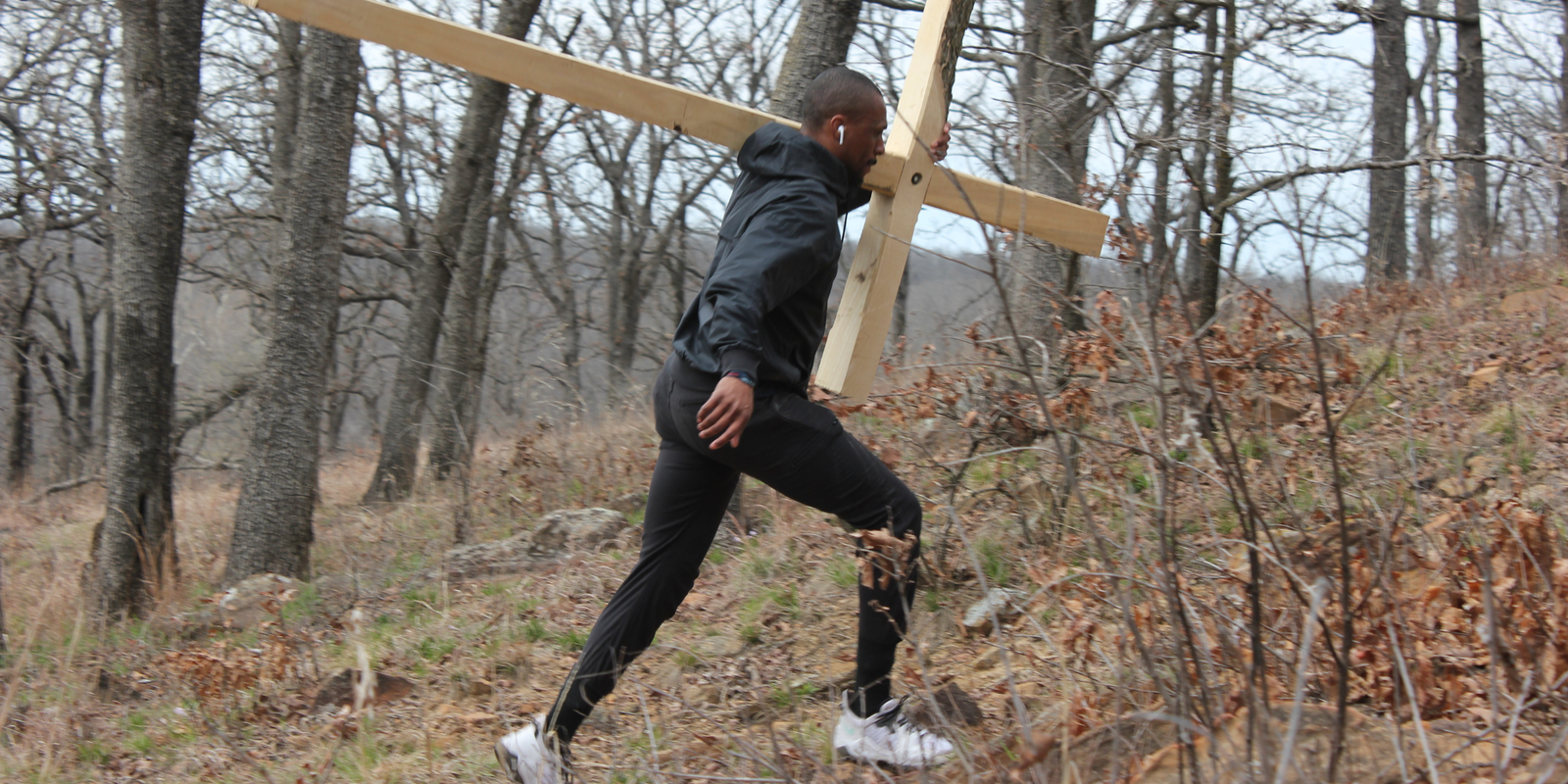 A person in athletic clothing is running through a forested area while carrying a large wooden cross on their back. The background features leafless trees and dry foliage.