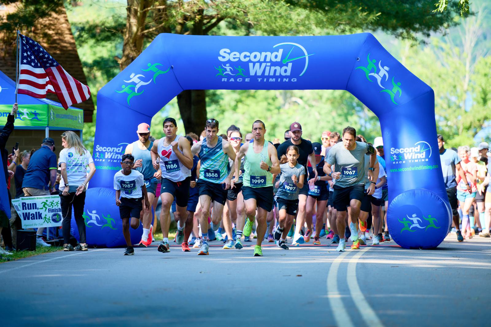A group of runners participating in a race begin their run under a blue "Second Wind Race Timing" inflatable arch. The event is set outdoors on a sunny day, with trees in the background and spectators on the sidelines.