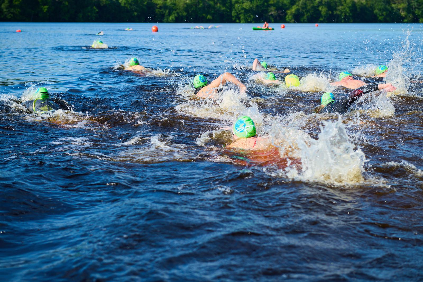A group of swimmers wearing green caps are racing in a lake. The water splashes around them as they swim towards brightly colored buoys on a sunny day. Lush trees line the background under a clear blue sky.