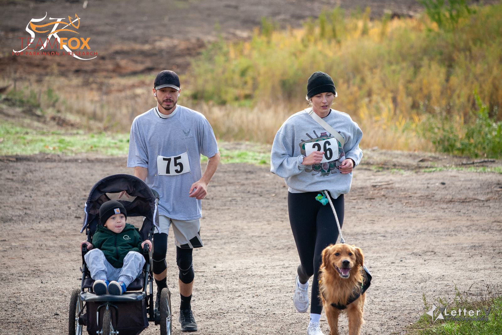 A man and a woman are jogging with a child in a stroller and a golden retriever on a leash. The woman wears a grey sweatshirt and black leggings, and the man wears a white shirt and black shorts. Both have race numbers. The path is outdoors with grassy surroundings.