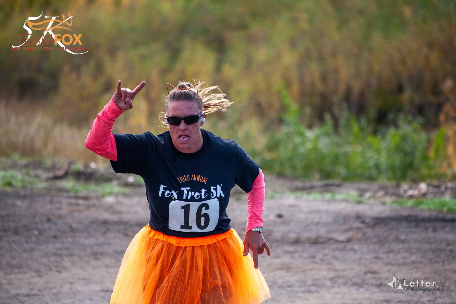 A person wearing a black "Fox Trot 5K" t-shirt and an orange tutu runs outdoors, making a hand gesture. They have sunglasses and cat ear headband, with tall grass in the background.