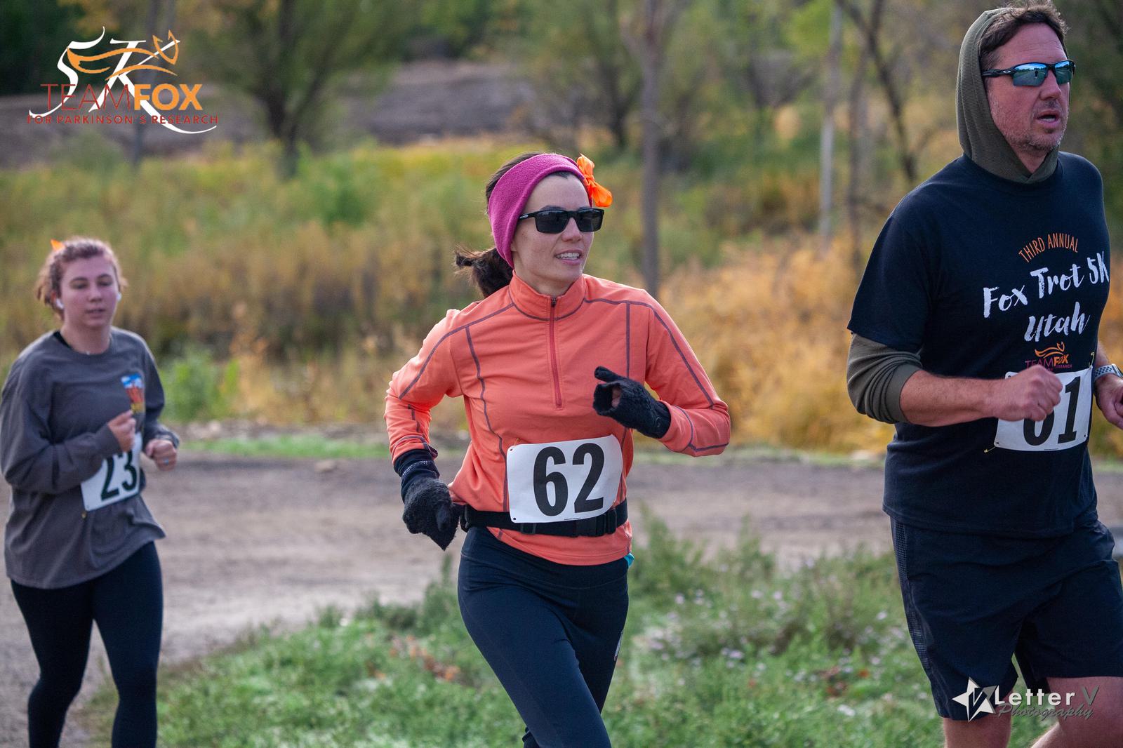Three people are running in an outdoor race. The person in the foreground wears a number 62 bib, an orange jacket, and a pink headband. Behind, two others wear black shirts with event branding. The background features grass and trees.
