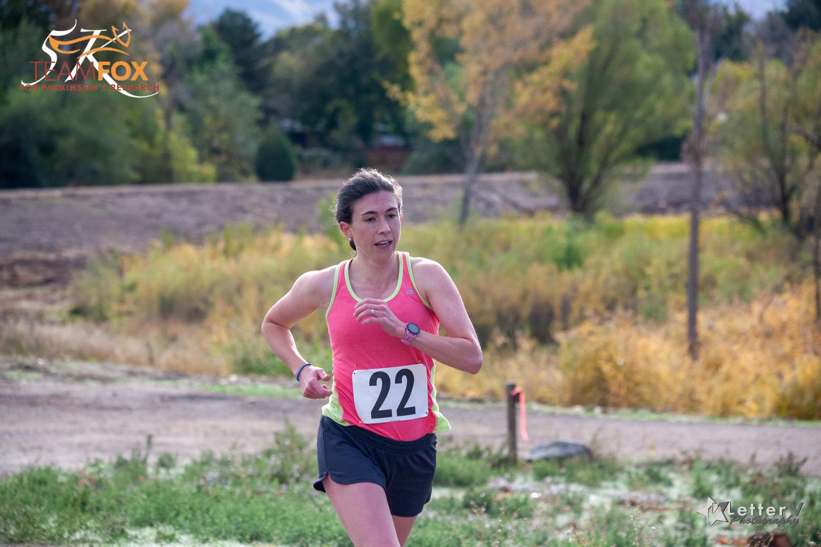 A person wearing a pink sleeveless top and black shorts is running with a number 22 bib. They are outdoors on a grassy path with a backdrop of trees and hills. A logo is in the top left corner of the image.