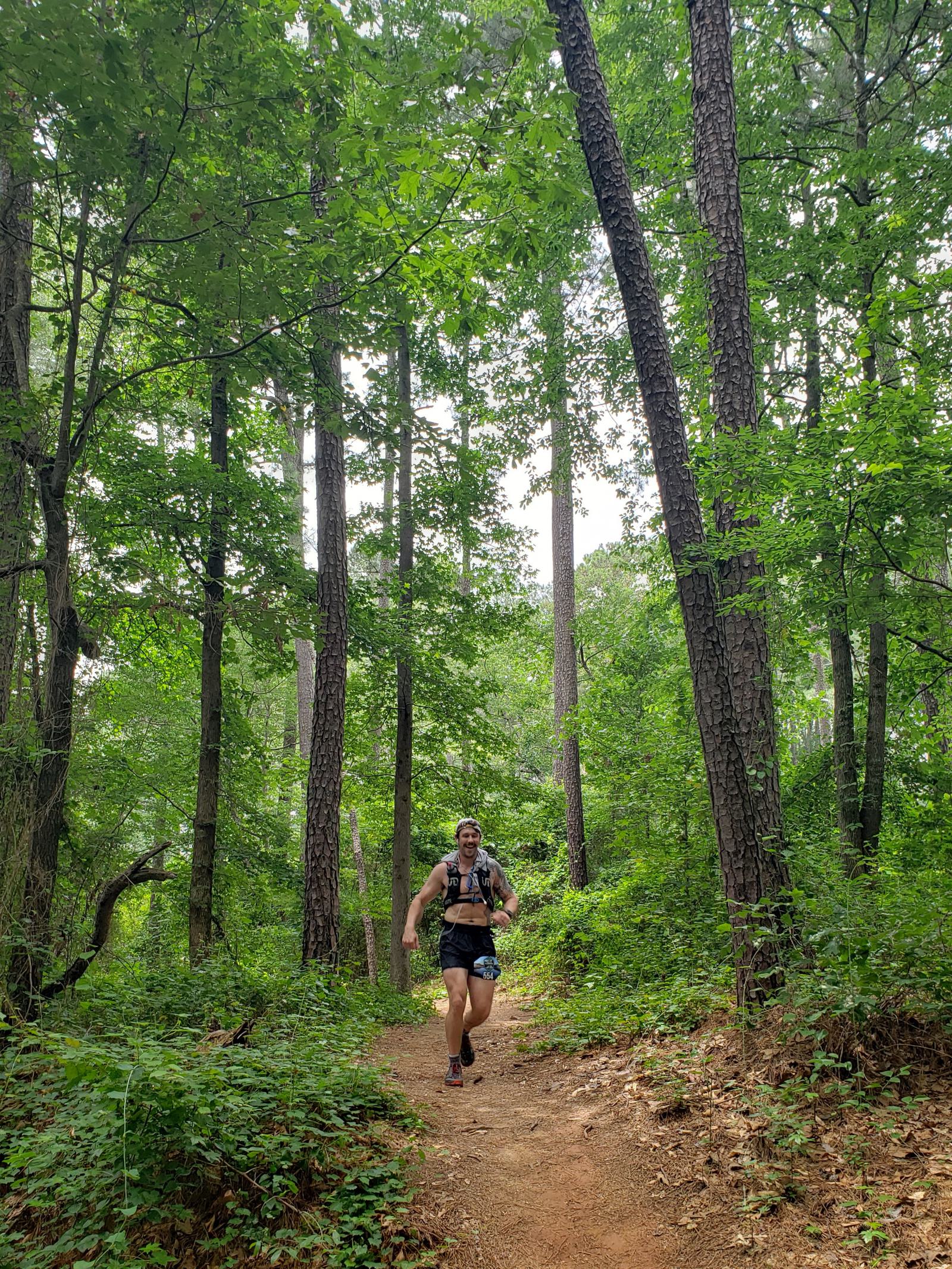 A person is running along a dirt trail through a lush green forest. Tall trees surround the path under a clouded sky, creating a peaceful and natural setting. The runner wears a backpack and athletic wear.