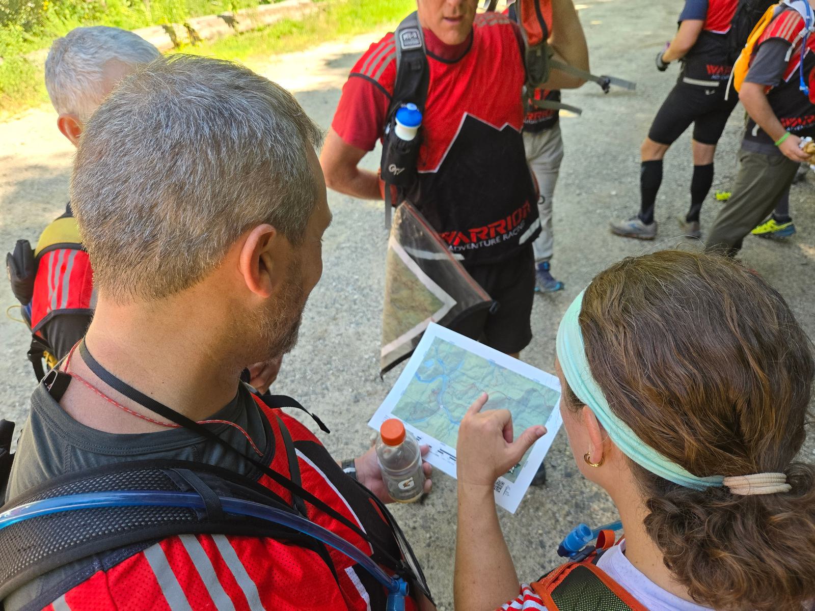 A group of people in red athletic shirts study a map outdoors; one person points at the map while others look on, preparing for an activity in a sunlit, wooded area.