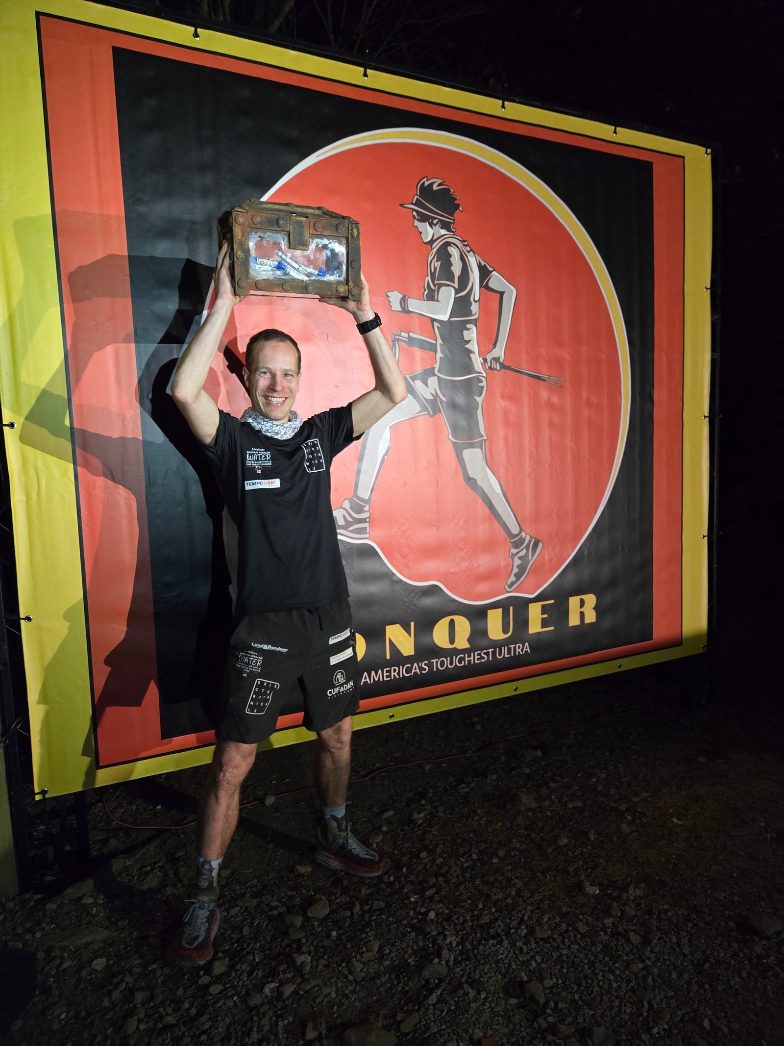 A person in athletic gear triumphantly holds a plaque overhead, standing in front of a large banner featuring a silhouette of a runner and the word "CONQUER." The scene appears to be outdoors at night.