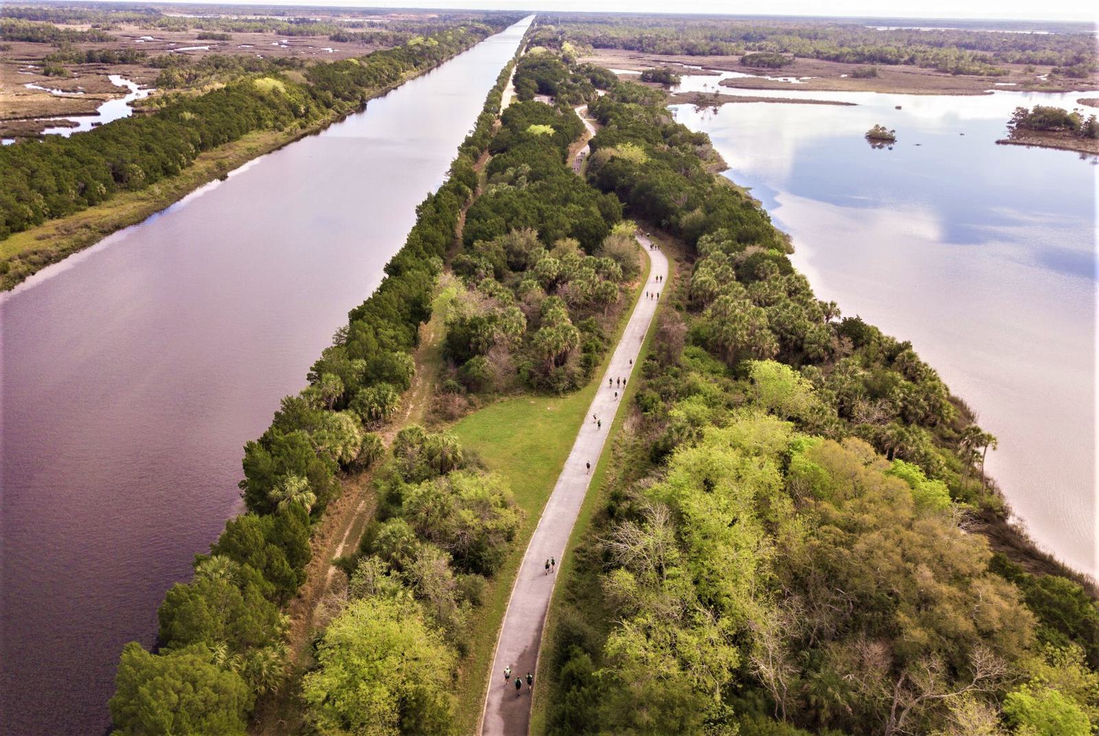 Aerial view of a scenic pathway flanked by lush greenery and two large bodies of water on either side. People can be seen walking along the path. The landscape extends toward the horizon with forests and fields in the distance.
