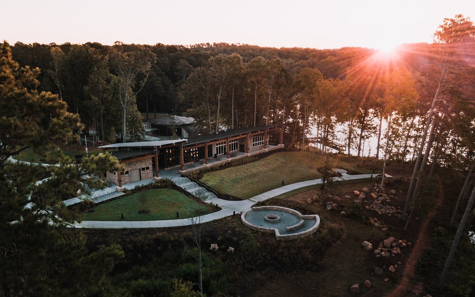 A modern building surrounded by trees sits beside a lake at sunset. The sun casts a warm glow over the landscape, illuminating the lush greenery and highlighting a circular path and grassy areas in the garden.