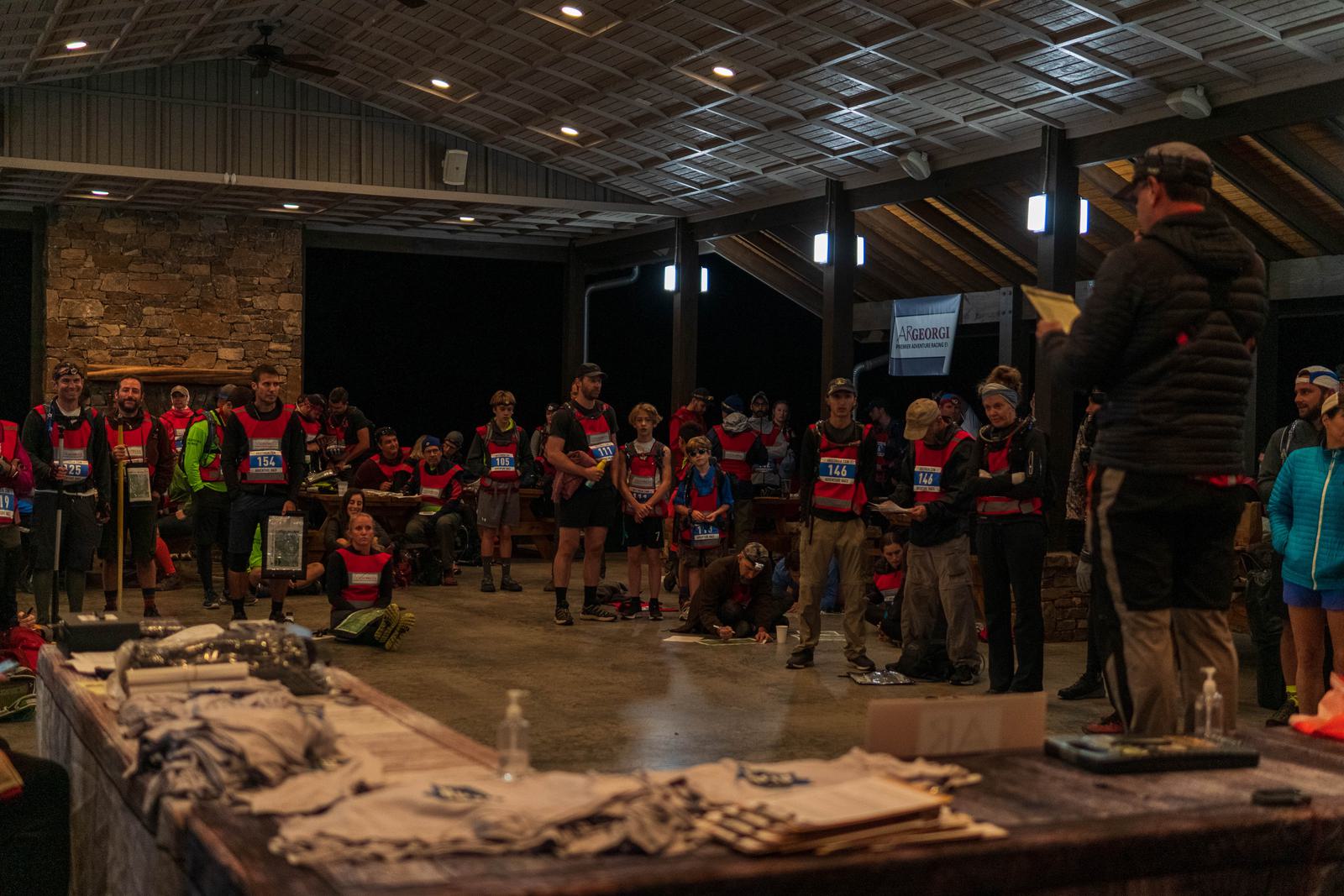 A group of runners wearing race bibs gather indoors, listening to a speaker holding a paper. Tables with race materials are in the foreground. The atmosphere is focused, and the area is lit with warm lighting.
