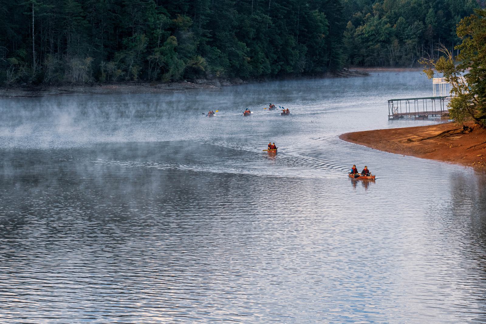 People paddle canoes and kayaks on a calm, misty lake surrounded by dense trees. A dock is visible on the right near a reddish-brown shoreline. The early morning light and fog create a tranquil scene.