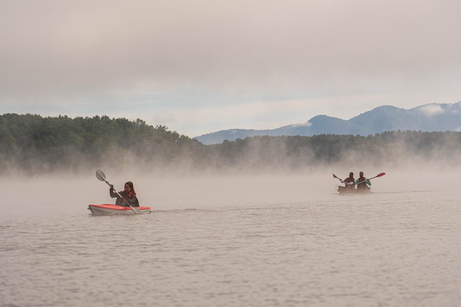 Three people kayaking on a calm, misty lake surrounded by trees, with distant mountains in the background under a cloudy sky.