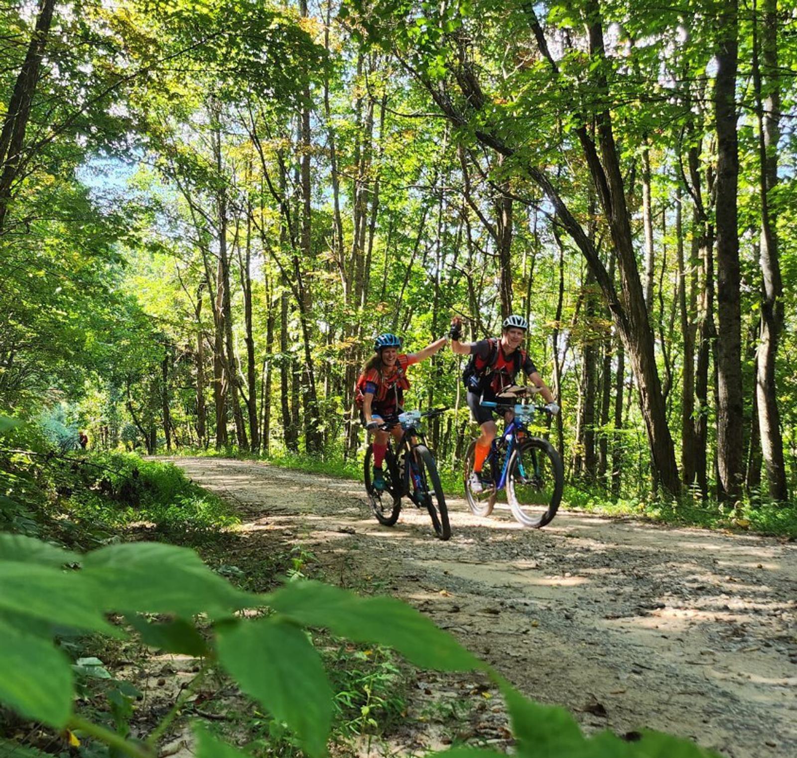 Two people wearing helmets ride bicycles on a dirt path through a lush green forest. One cyclist playfully reaches out to the other as sunlight filters through the tall trees overhead.
