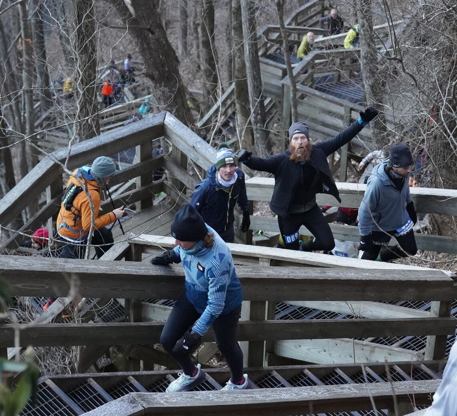 Runners ascend steep wooden stairs on a forest trail. Some look determined, while one man in black spreads his arms wide, appearing joyful. The scene is energetic, with leafless trees in the background.