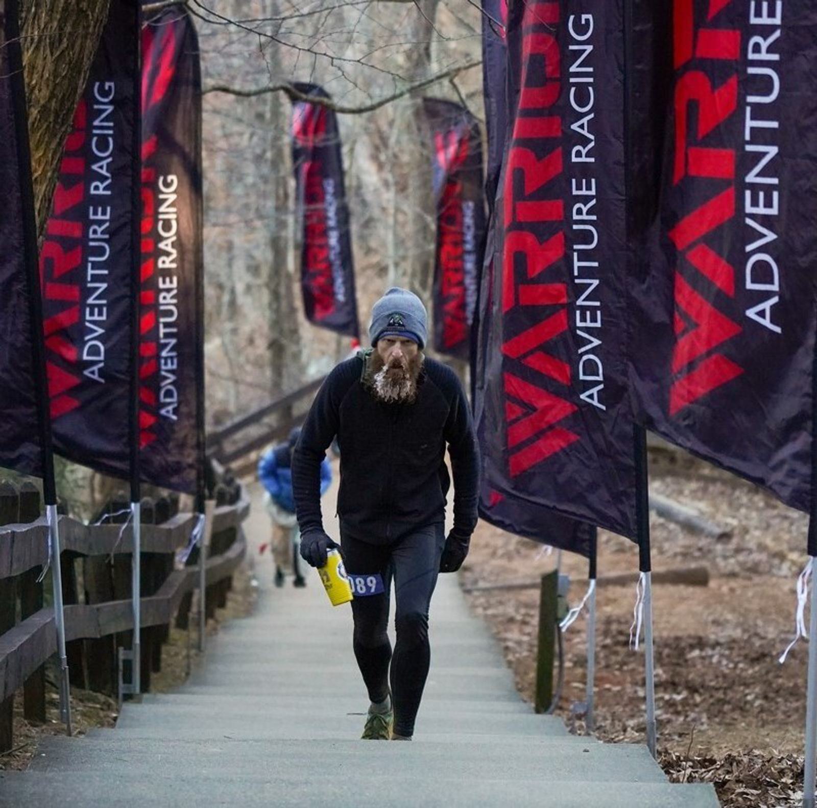 A bearded man in athletic gear and a beanie climbs outdoor steps during a race, holding a yellow water bottle. Black and red banners reading "WARRIOR ADVENTURE RACING" line the path in a wooded area.
