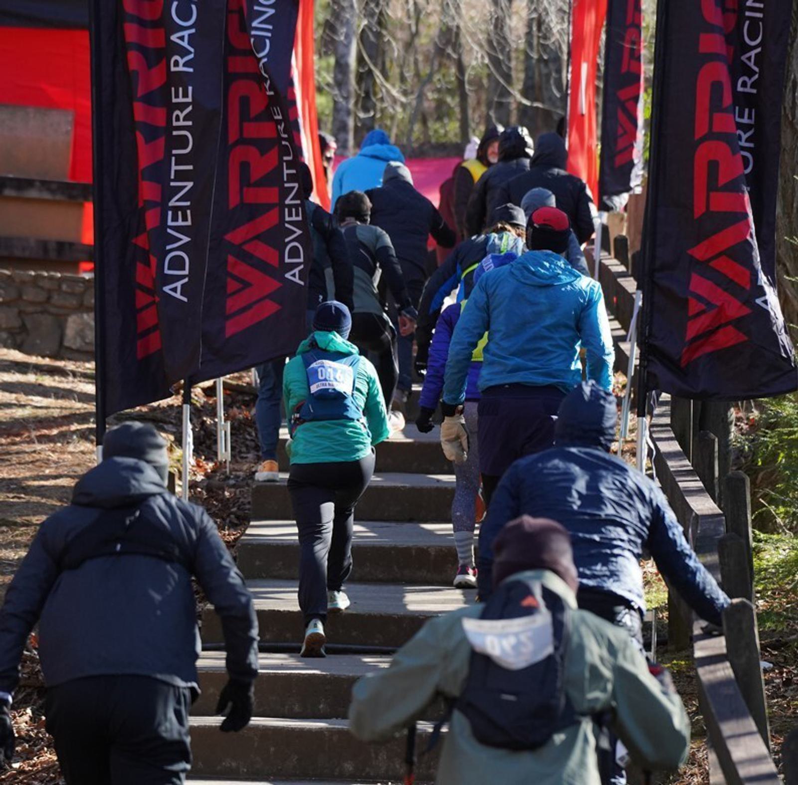 A group of runners wearing winter gear climb outdoor stairs during an adventure race, flanked by black and red "WARRIOR ADVENTURE RACING" banners. The scene is set in a wooded area on a sunny day.