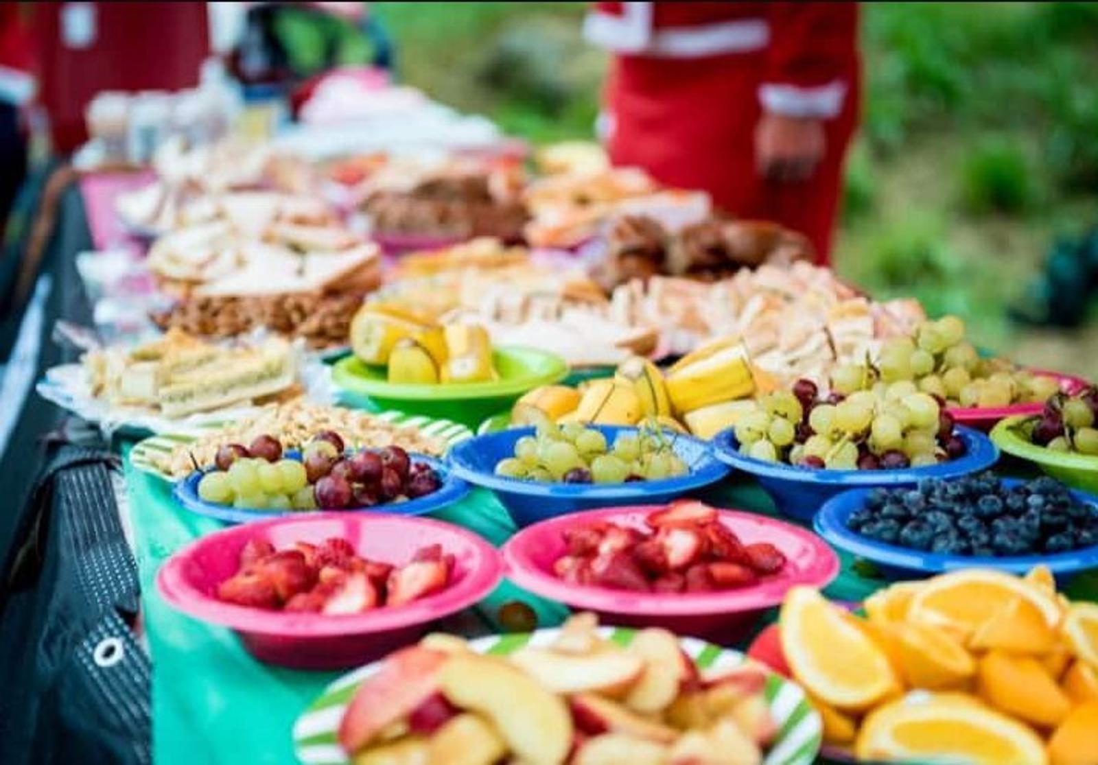 A colorful assortment of fruits and snacks arranged on a long table. There are plates of grapes, strawberries, oranges, blueberries, and sliced sandwiches. The table is covered with a green cloth, with people blurred in the background.
