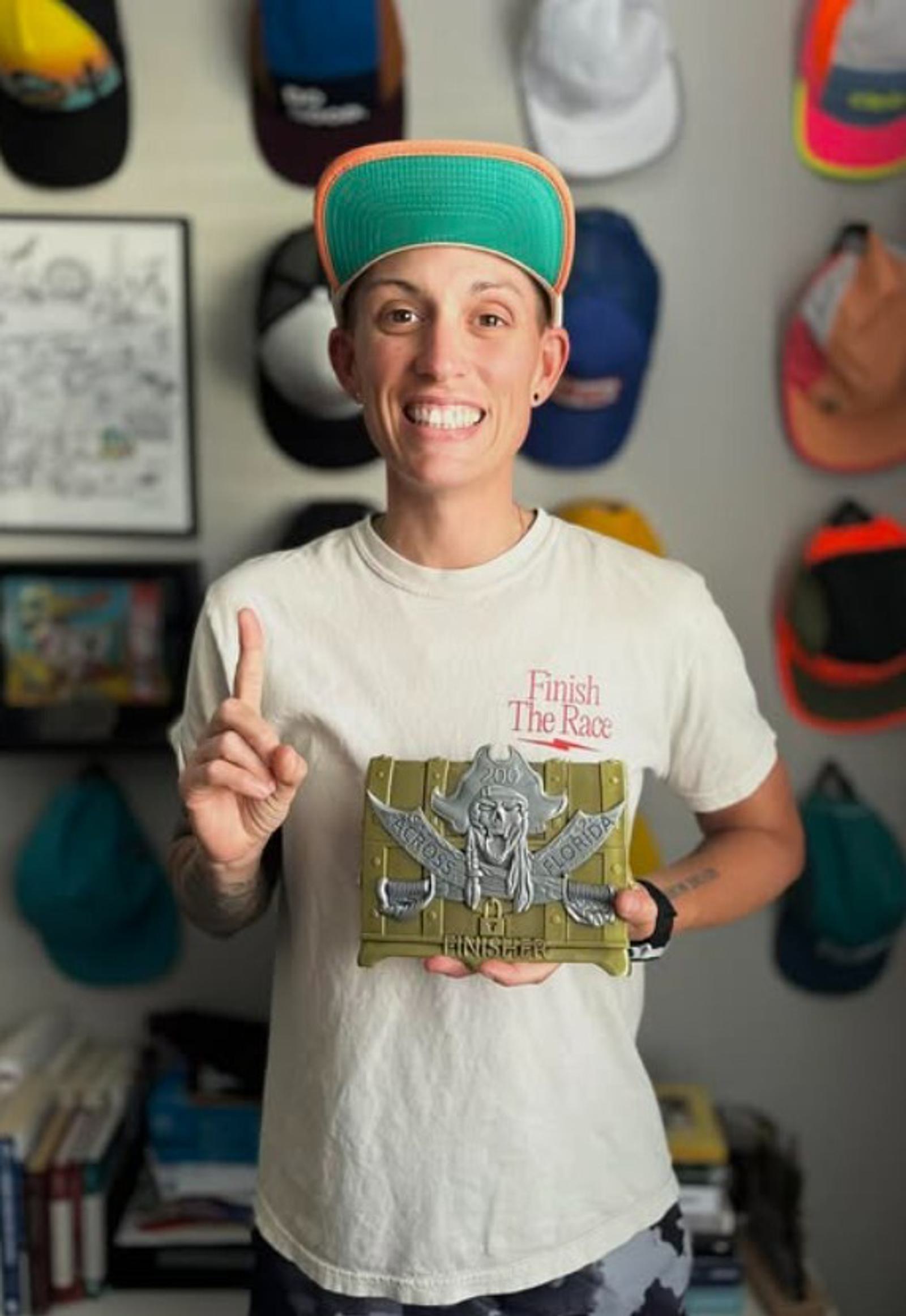 A person wearing a white "Finish The Race" t-shirt and a teal cap smiles while holding a trophy. They are standing in front of a wall with various hats displayed. Their right hand holds up one finger.
