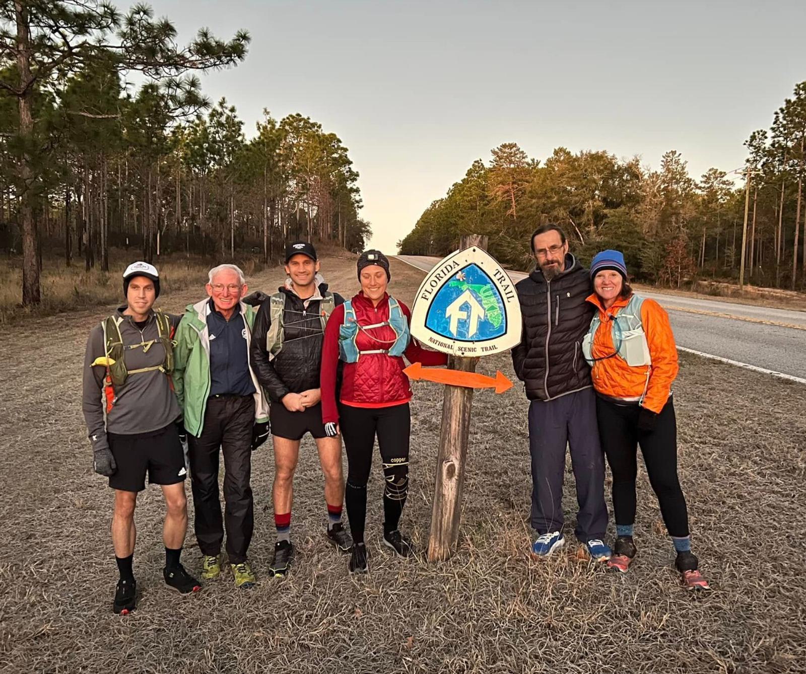 A group of six people dressed in hiking attire stand together on a grassy path beside a road. They pose next to a wooden sign with a blue and white trail symbol, surrounded by trees under a clear sky.