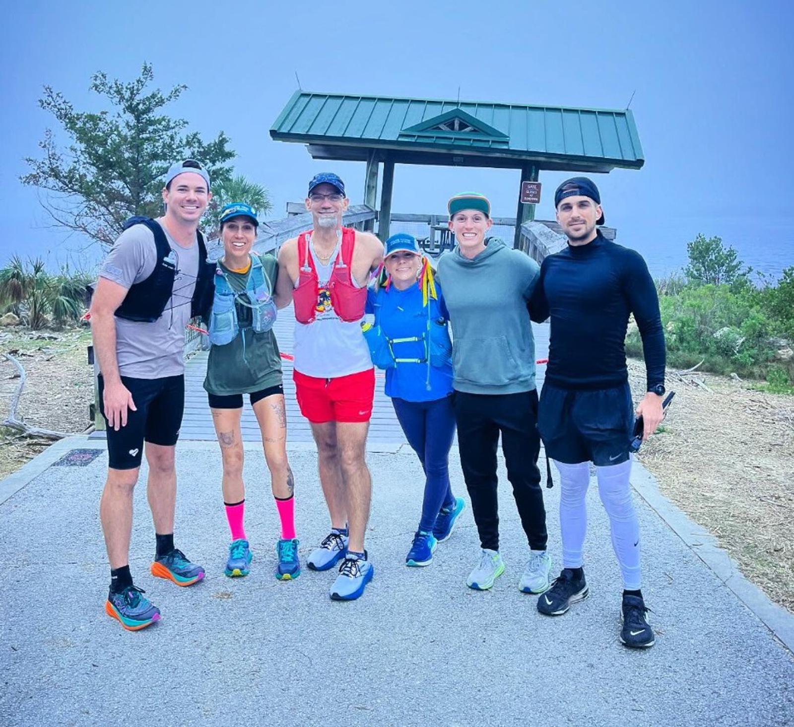 A group of six people wearing athletic gear poses together outdoors. They stand on a paved path with trees and bushes in the background. A pavilion with a green roof is visible behind them. The sky is overcast.