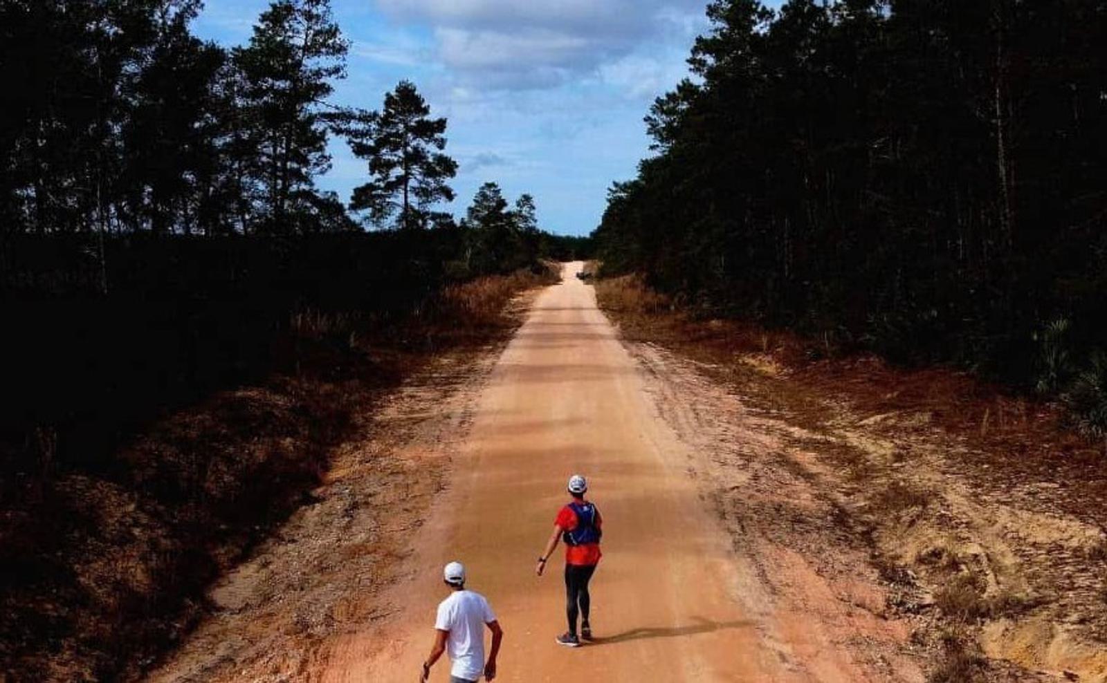 Two people walking on a wide dirt road lined with trees under a partly cloudy sky. One wears a white shirt, the other a red one with a blue backpack. The road stretches into the distance, surrounded by forest.