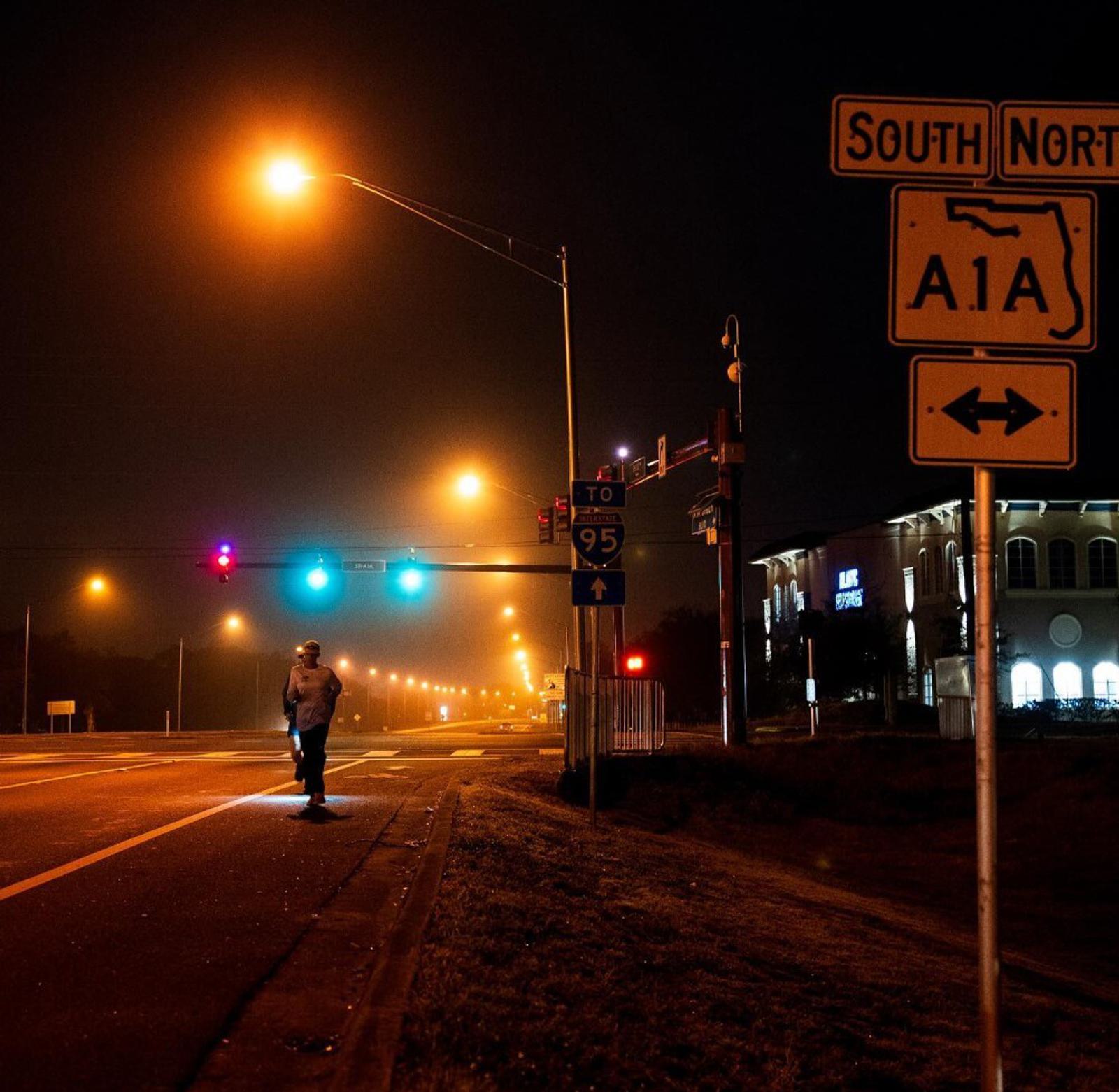 A solitary person walks along a dimly lit street at night. Streetlights cast an orange glow over the road. Traffic signals show red and green lights. Signs indicate directions for South North A1A and Interstate 95. A building is visible in the distance.