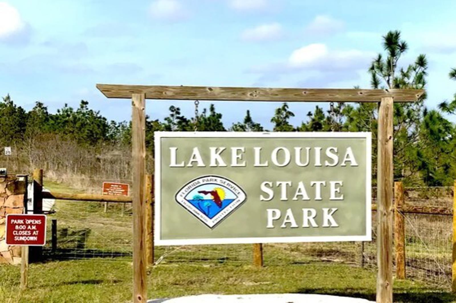 A wooden sign at the entrance of Lake Louisa State Park stands in front of grassy fields and pine trees. A smaller sign nearby notes park hours and closing time at sundown. The sky is partly cloudy.