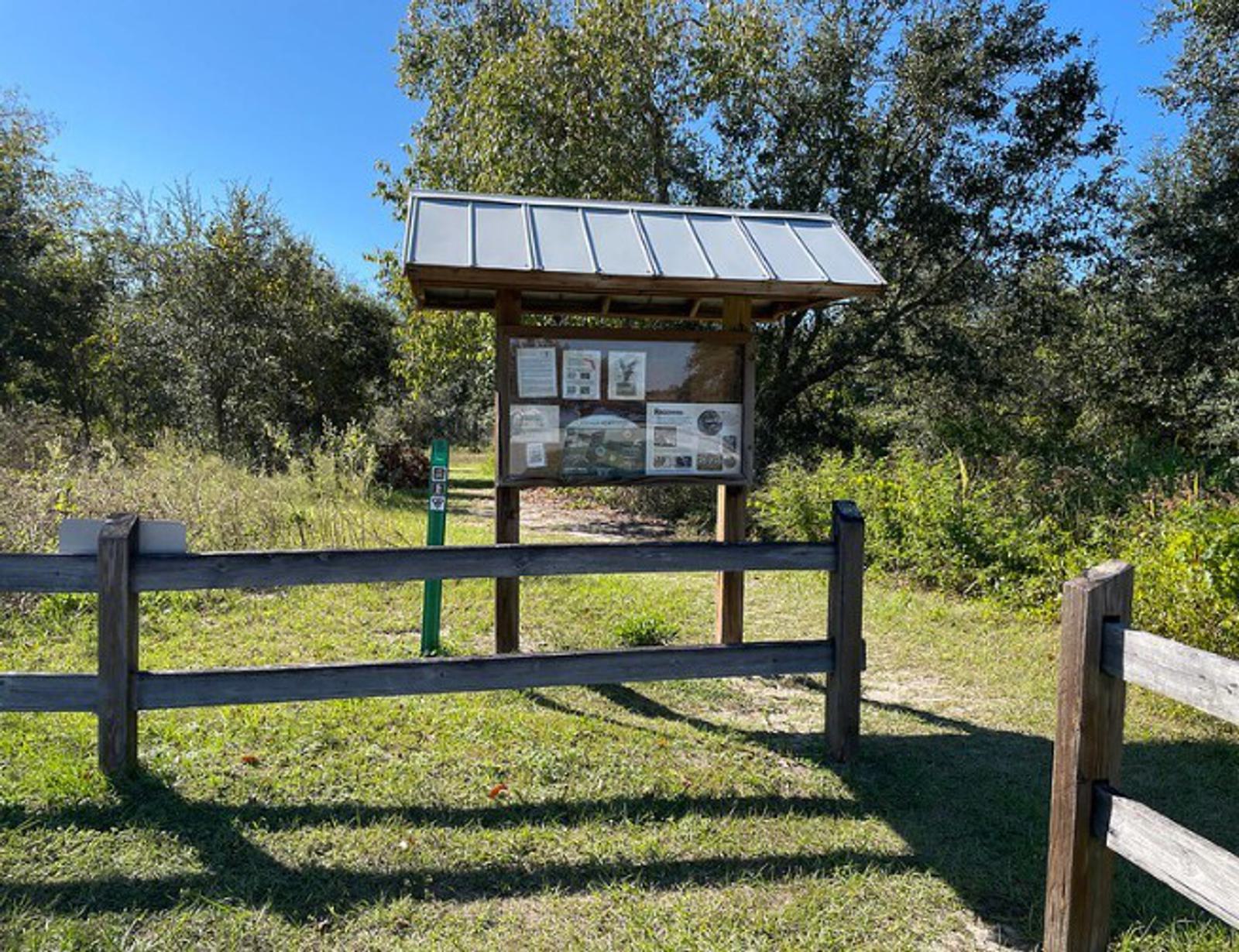A wooden information board with posted signs stands behind a split-rail fence at the entrance to a nature trail, surrounded by green grass, trees, and sunlight.