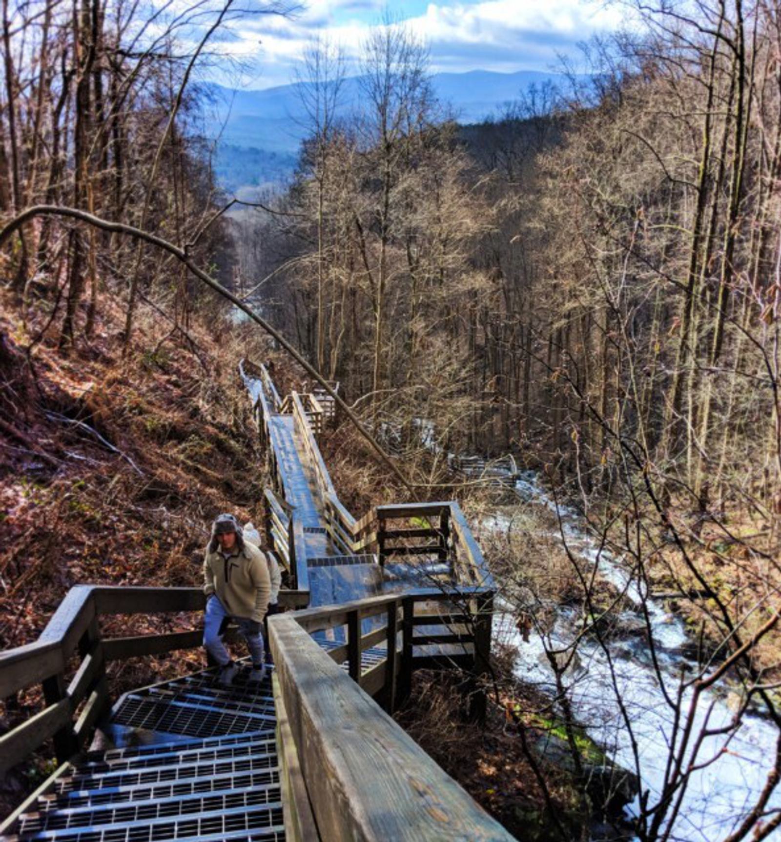 A person with a backpack climbs wooden stairs beside a waterfall in a forested, mountainous area on a clear day. Leafless trees line the path, and distant mountains are visible under a partly cloudy sky.