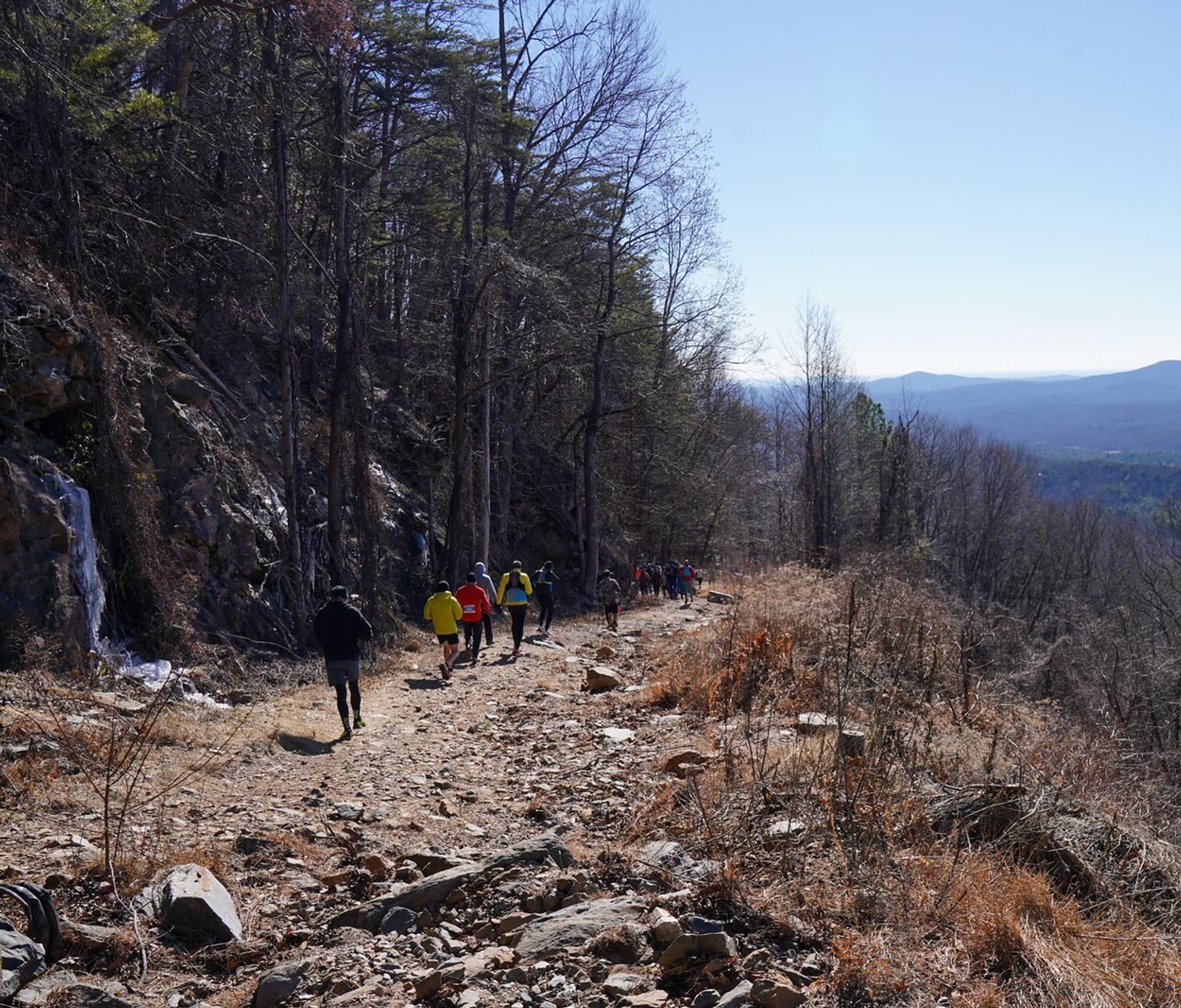 A group of people hike along a rocky mountain trail bordered by trees, with distant hills and a clear blue sky visible in the background.