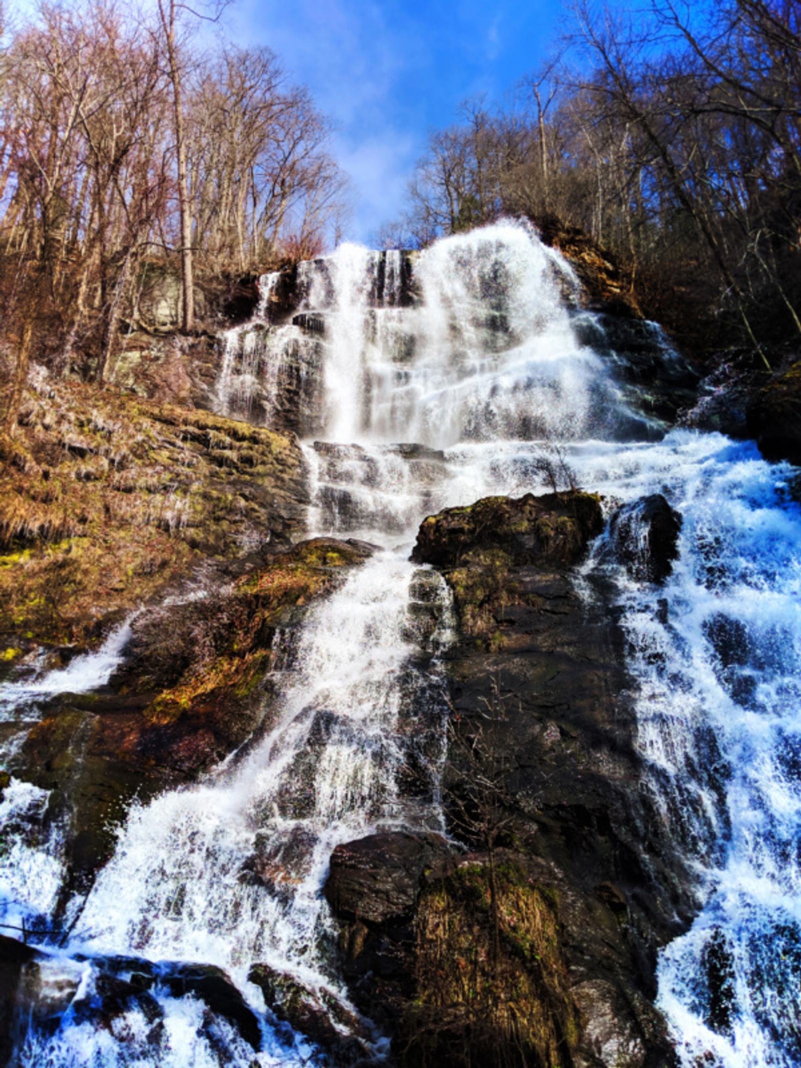 A tall waterfall cascades down a rocky cliff surrounded by bare trees under a clear blue sky. The water flows over multiple tiers, creating white foamy streams against the dark rocks. The scene is bright and vibrant.