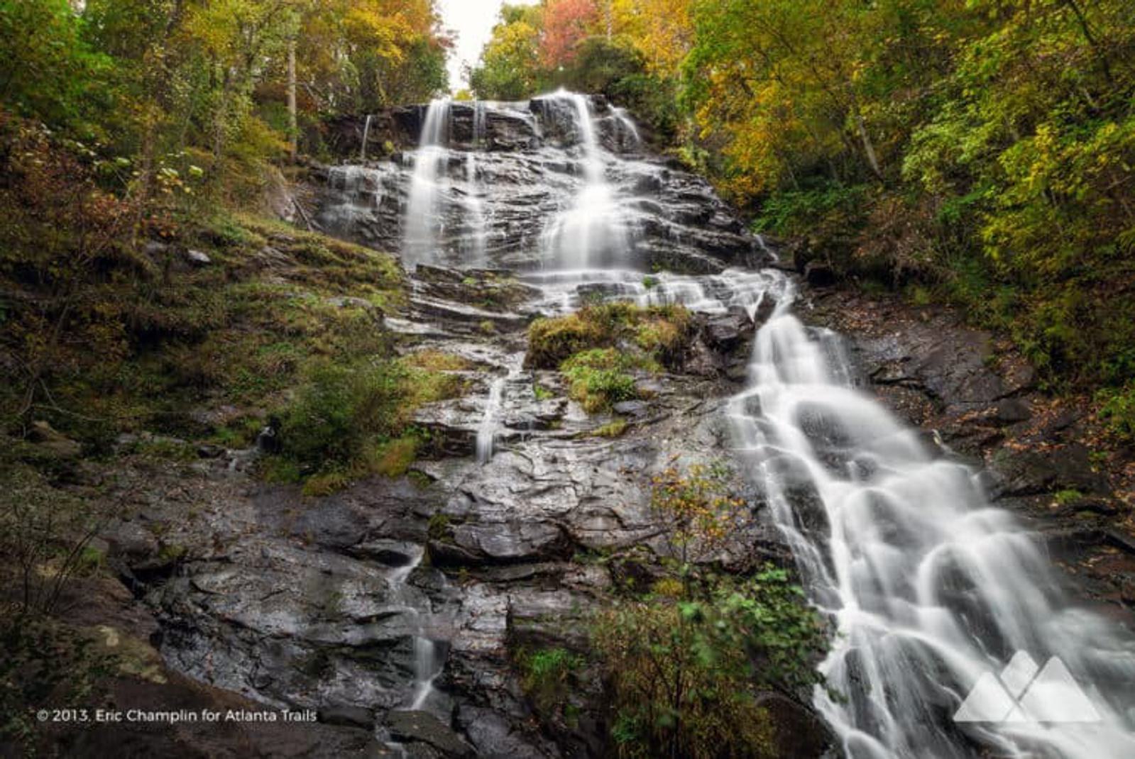 A wide, multi-tiered waterfall cascades over dark rocks surrounded by lush green, yellow, and orange foliage, suggesting an autumn forest scene. The water gently flows down the rocky surface.