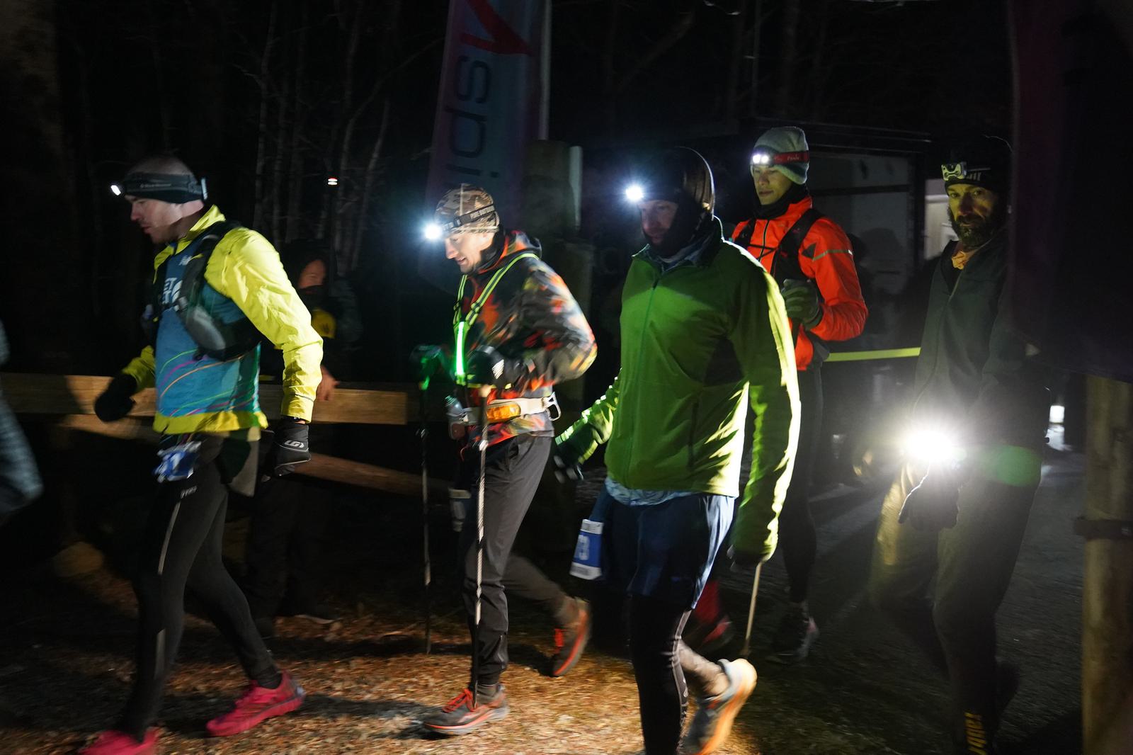 A group of runners wearing headlamps and winter gear competes in a nighttime race. They are on a dimly lit outdoor path, some carrying hydration packs and bottles. The scene is illuminated by their headlamps and a nearby light source.
