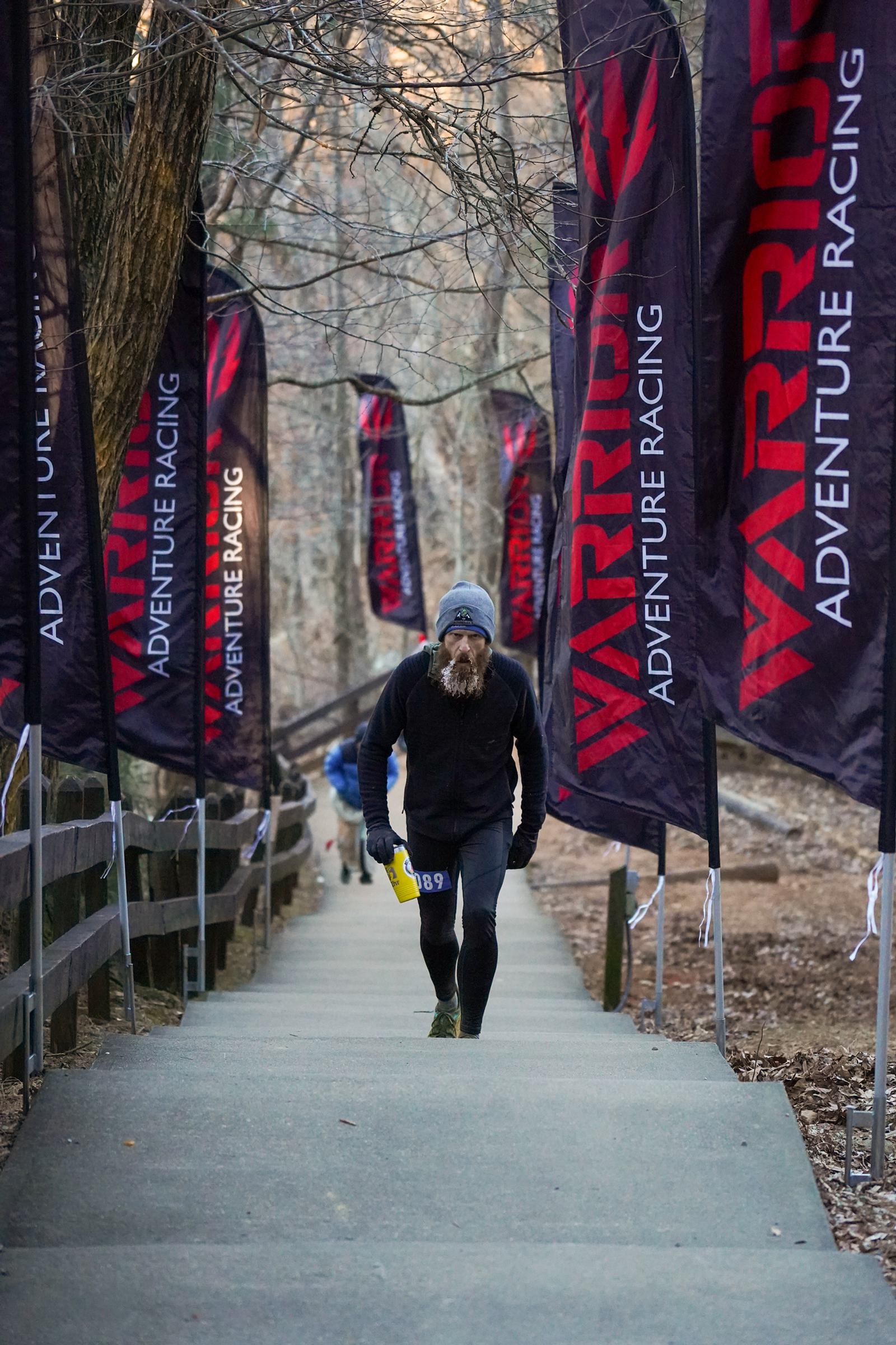 A person wearing athletic gear and a beanie climbs outdoor stone steps lined with "Warrior Adventure Racing" flags in a wooded area. The steps are surrounded by trees, and sunlight filters through the branches.