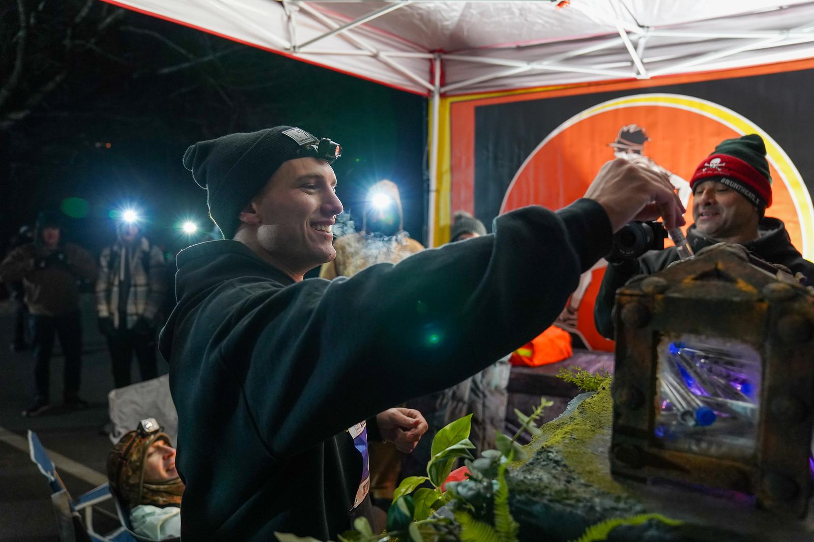 A person in a black hoodie and beanie smiles while reaching towards a small, ornate box. They are surrounded by others under a canopy, with bright lights in the background. The atmosphere appears festive and lively.