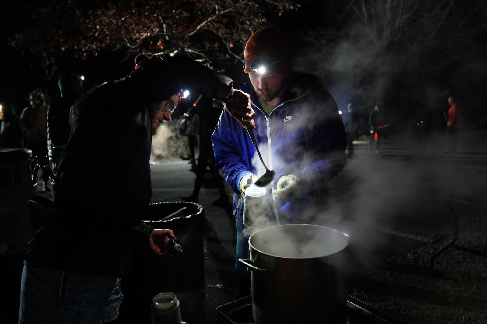 Two people wearing headlamps serve steaming hot food from a large pot outdoors at night. Both are dressed warmly, indicating a cold setting. The scene is dimly lit, with other figures visible in the dark background.