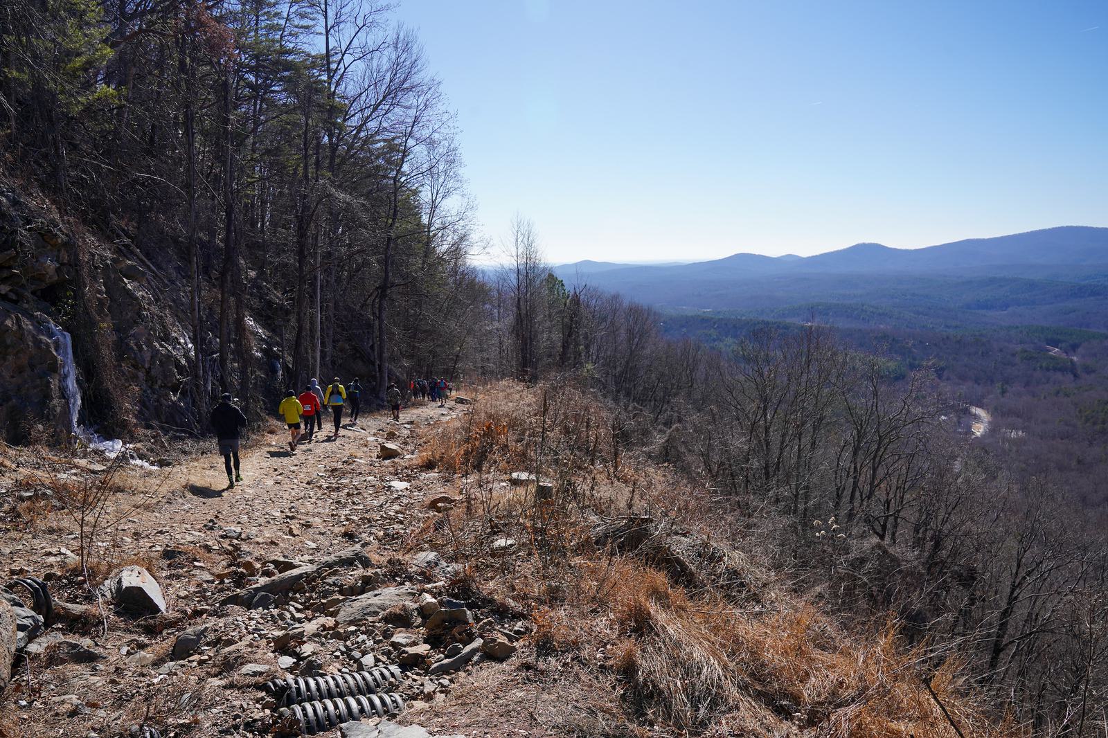 A group of hikers walk along a rocky mountain trail bordered by trees, with a scenic view of distant hills and valleys under a clear blue sky.