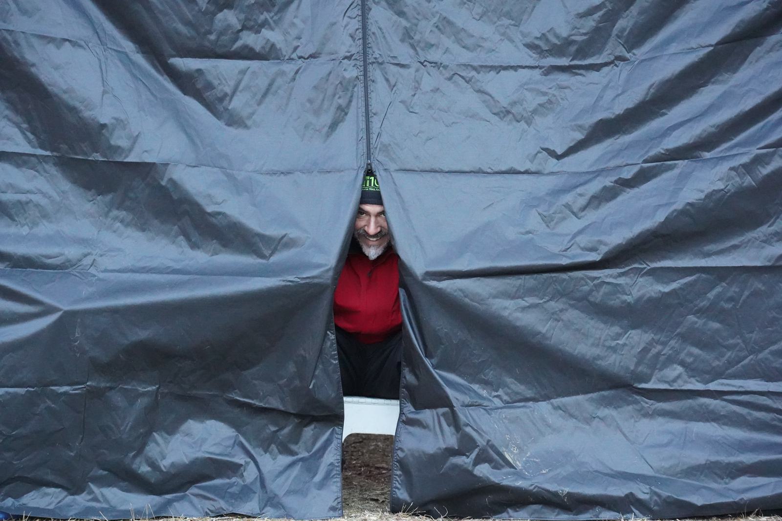 A person wearing a red jacket and a dark beanie peeks out from behind the gap in a large gray sheet or tarp. The surroundings suggest an outdoor setting.