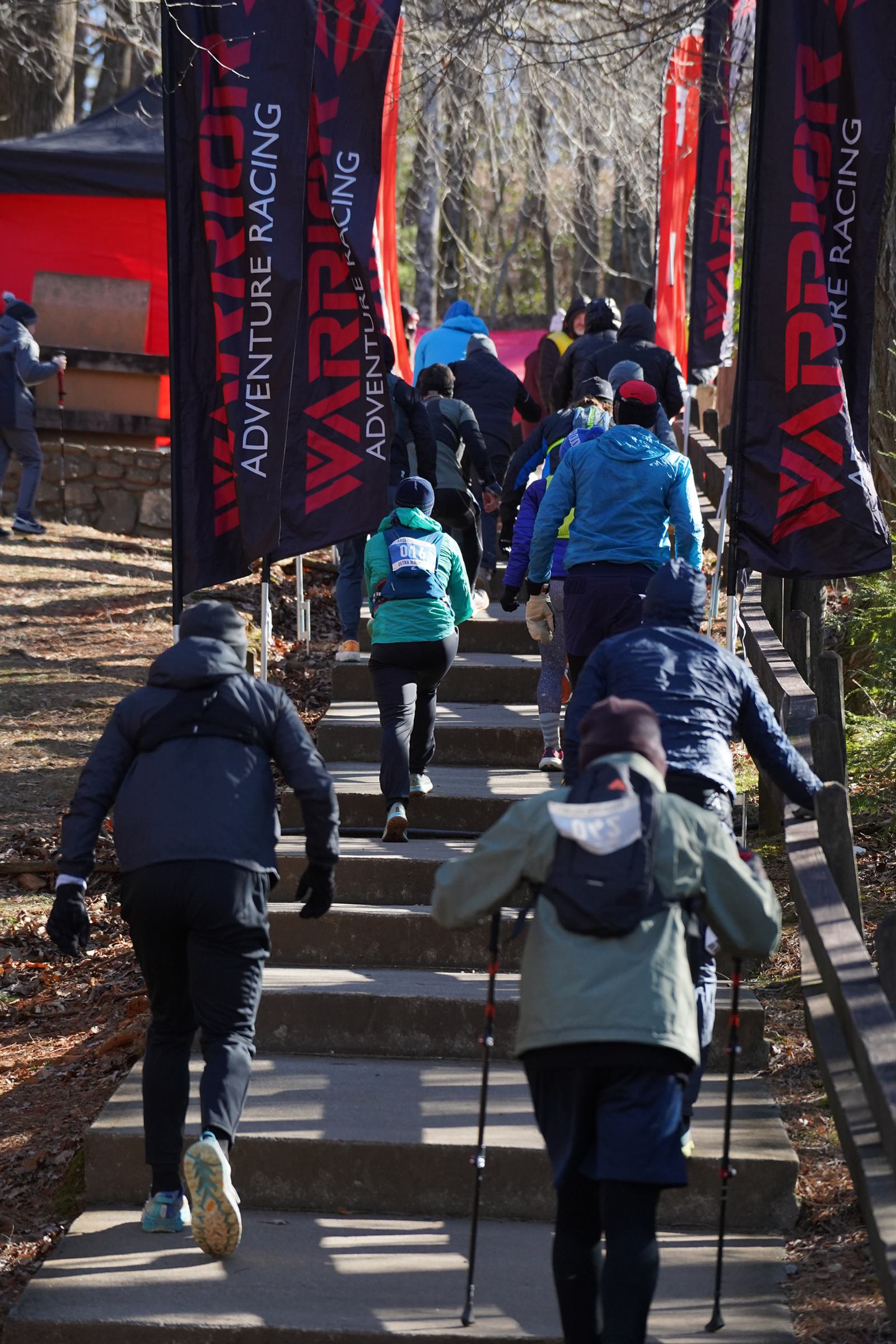 Participants in outdoor gear ascend concrete steps during the Warrior Adventure Racing event. Banners with "Warrior Adventure Racing" flank the path, and trees are visible in the background. Some people use trekking poles.