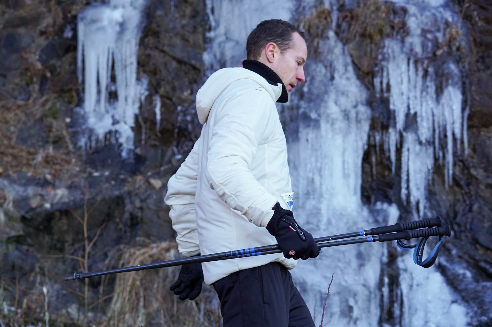 A person in a white jacket and black gloves walks past a rocky, frozen waterfall, holding trekking poles. The icy formations and bare rock suggest a cold, winter environment.