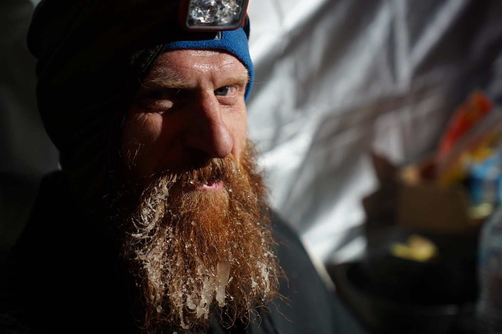 A person with a frost-covered beard and a headlamp sits indoors, partially in shadow. They wear a blue head covering and dark clothing. The background is slightly blurred but shows indistinct objects and warm lighting, suggesting an enclosed space.