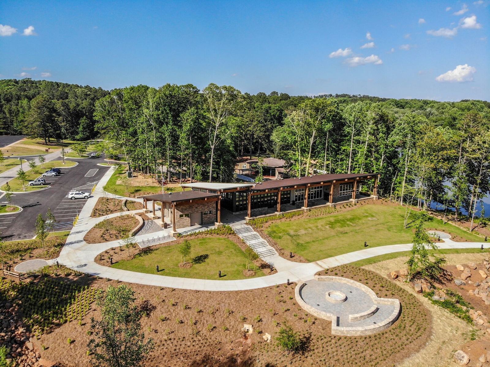 Aerial view of a modern building surrounded by lush forest. The structure features large glass windows and a flat roof. A circular pathway and landscaped garden are in the foreground. A parking lot with several cars is visible to the left.