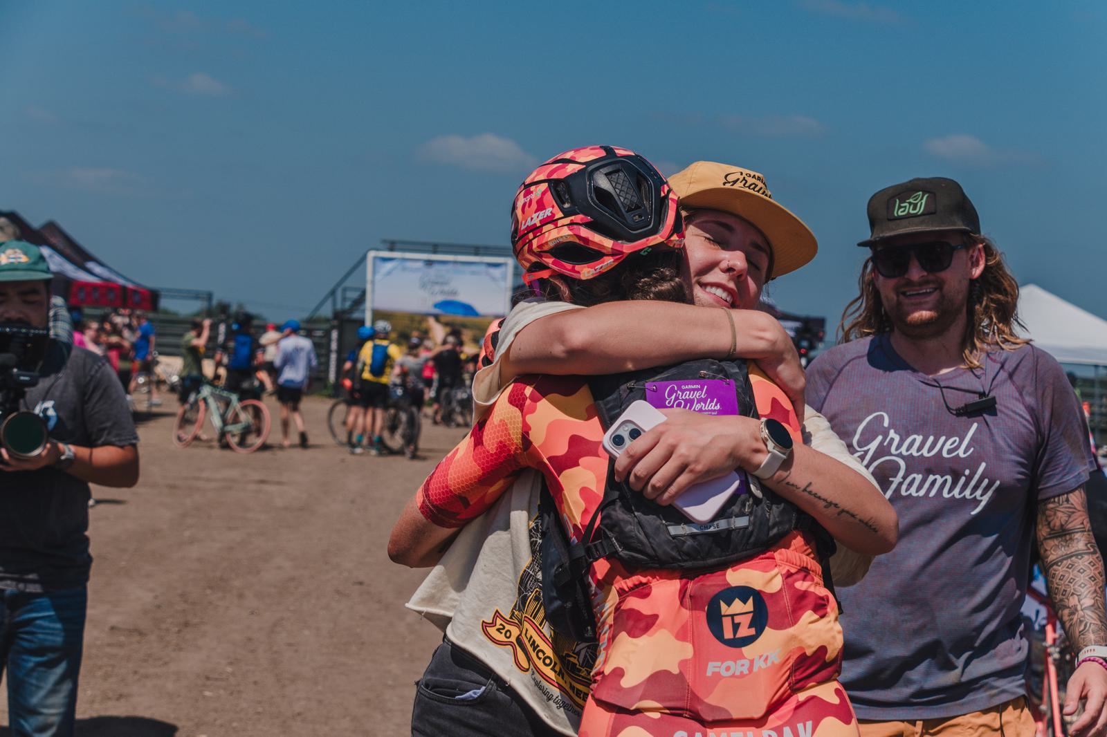 Two cyclists in helmets and casual clothing share a joyful hug in a sunny outdoor setting. One holds a phone, and another person wearing sunglasses and a "Gravel Family" shirt stands nearby. Bicycles and a camera operator are visible in the background.