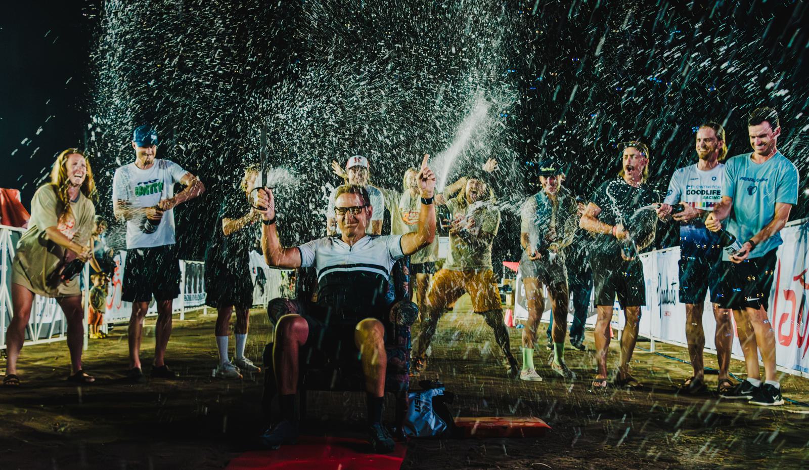 A group of people celebrate outdoors at night, spraying champagne. A person seated in the front raises both arms triumphantly while others cheer around them. Water splashes and excitement fill the scene.