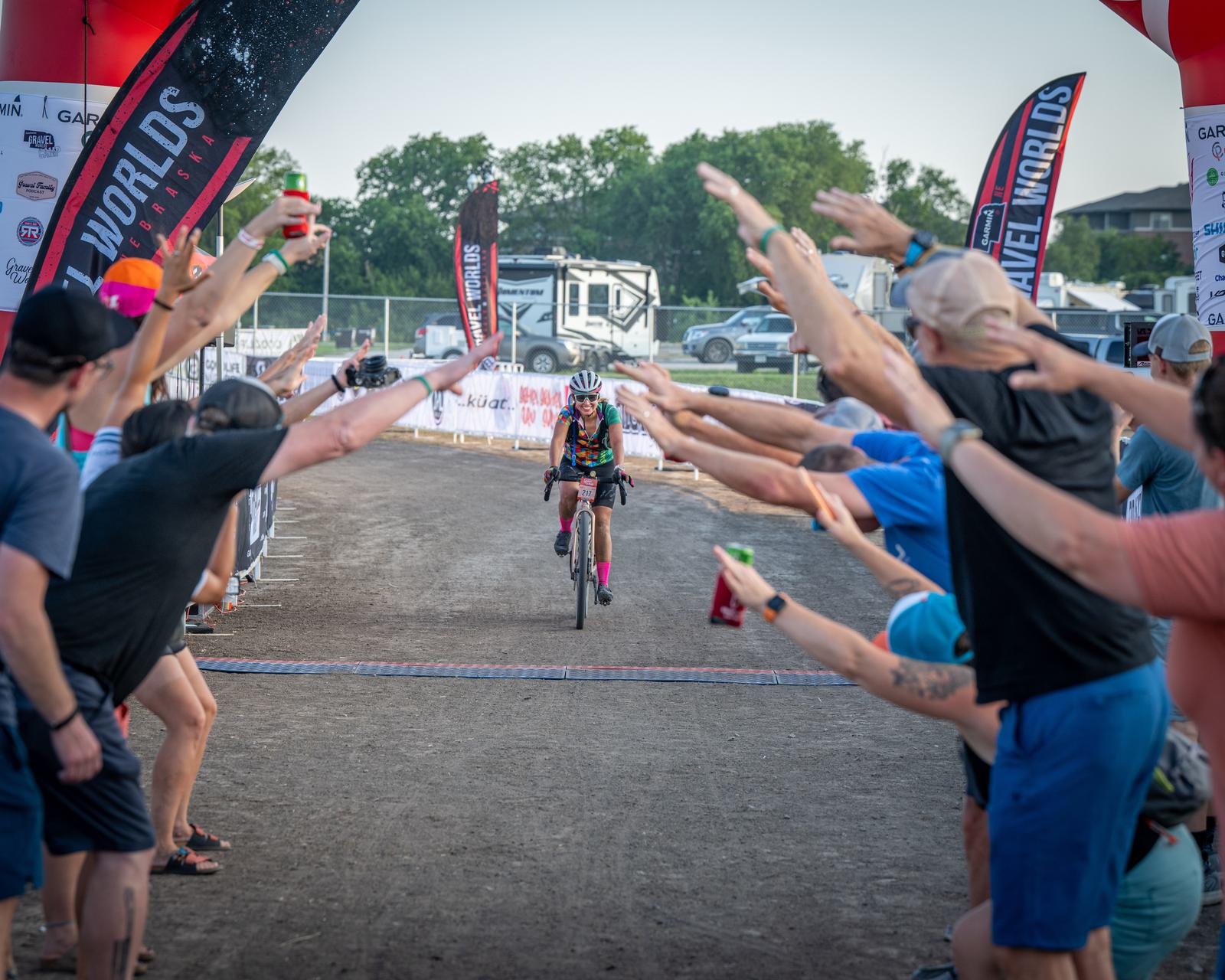 A cyclist reaches the finish line at an outdoor race, greeted by cheering spectators forming a tunnel with their arms. The ground is dirt, and there are banners and flags lining the path. The background features trees and parked vehicles.
