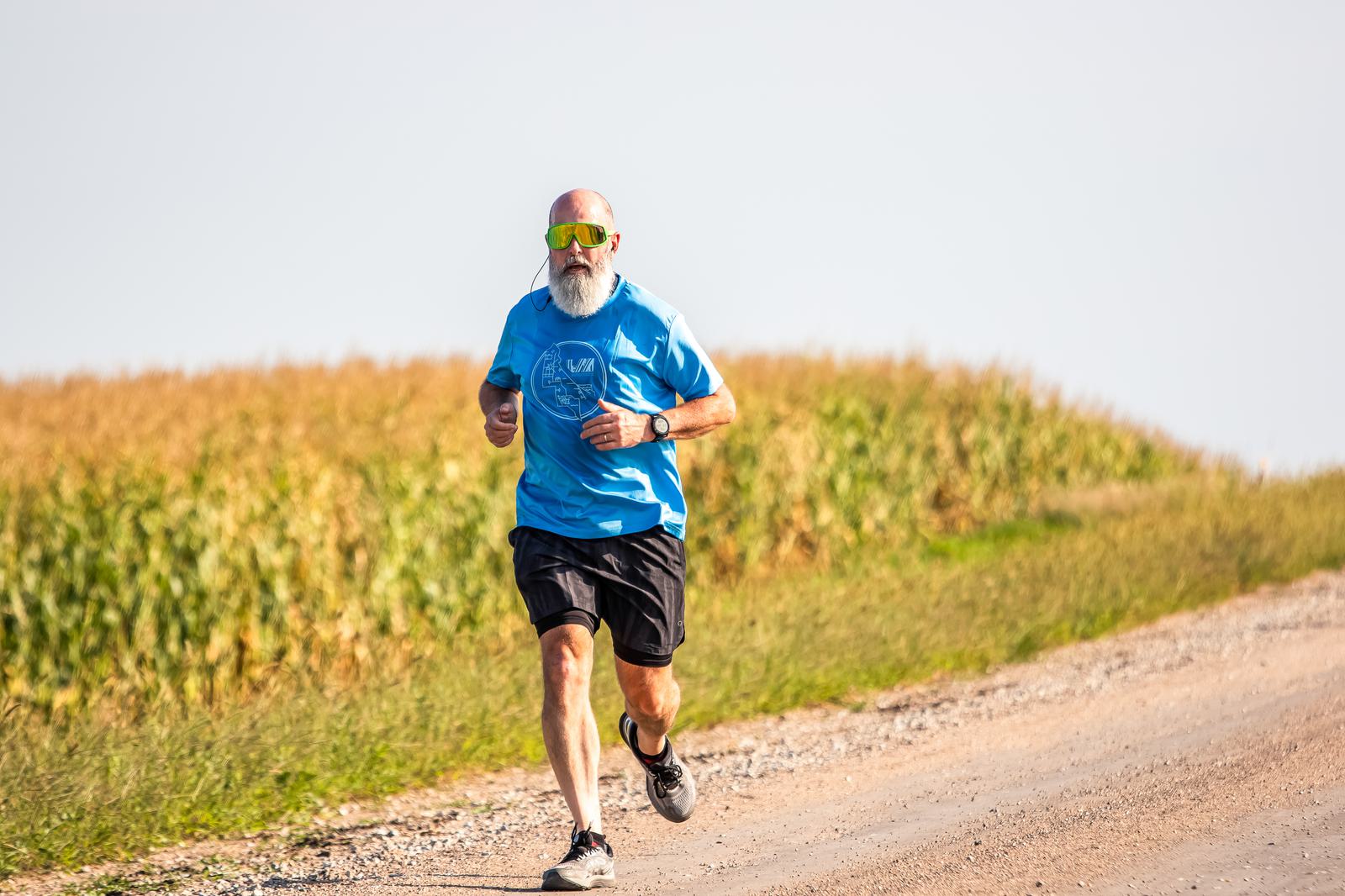 An older man with a white beard and sunglasses is running on a gravel path beside a cornfield. He is wearing a blue shirt, black shorts, and running shoes. The sky is clear, suggesting a bright, sunny day.