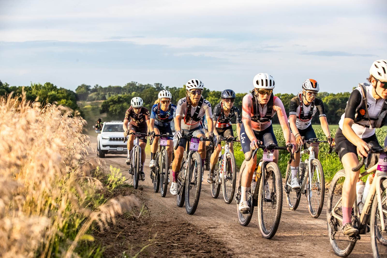 A group of cyclists in racing gear rides on a dirt road flanked by tall grasses. A white vehicle follows behind. The sky is clear with a few clouds, and the landscape is lush and green, suggesting a warm day.