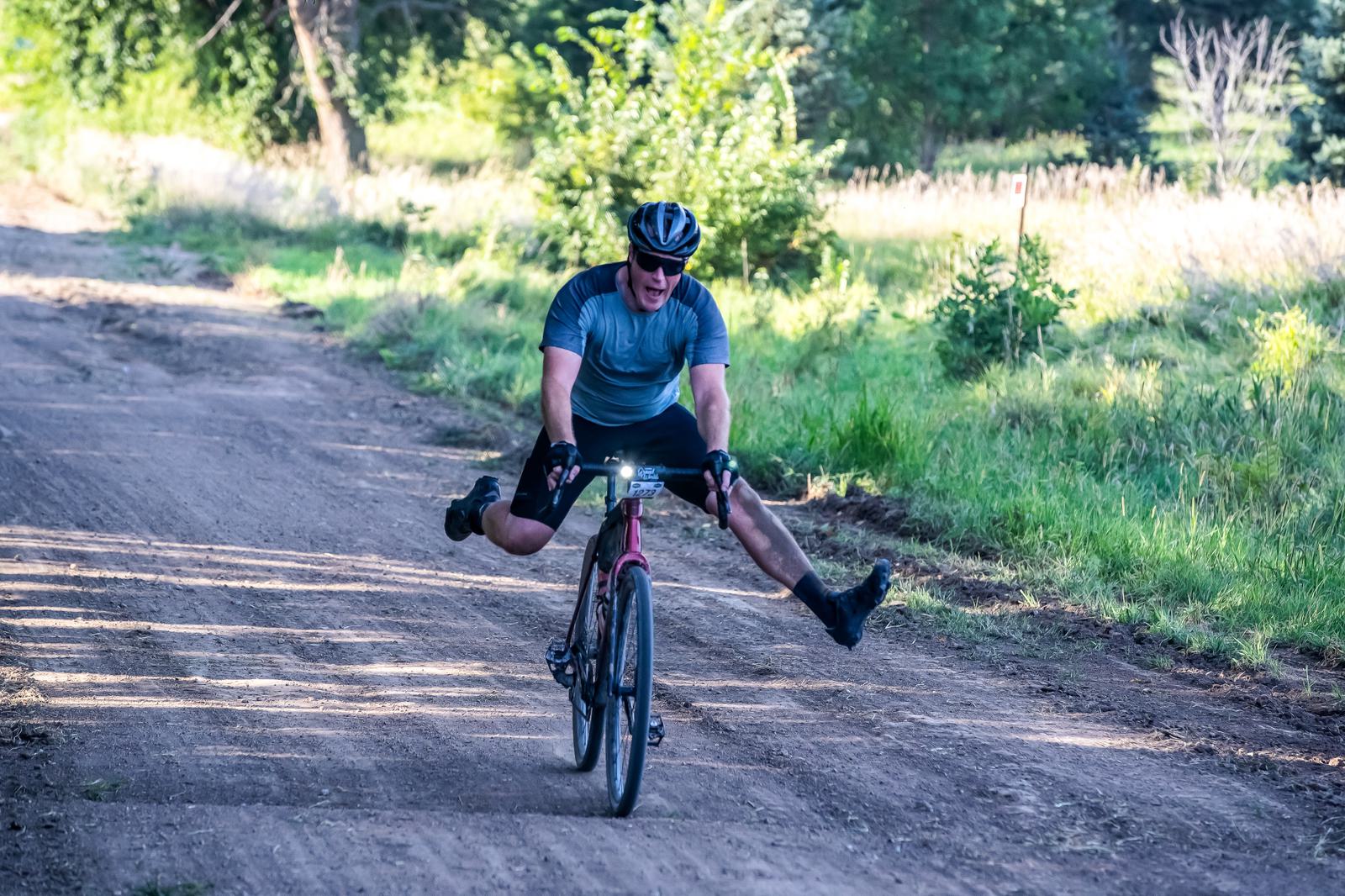 Person riding a bicycle on a dirt path with both legs extended out to the sides, wearing a helmet and blue shirt. Lush green trees and grass line the path under a sunny sky.