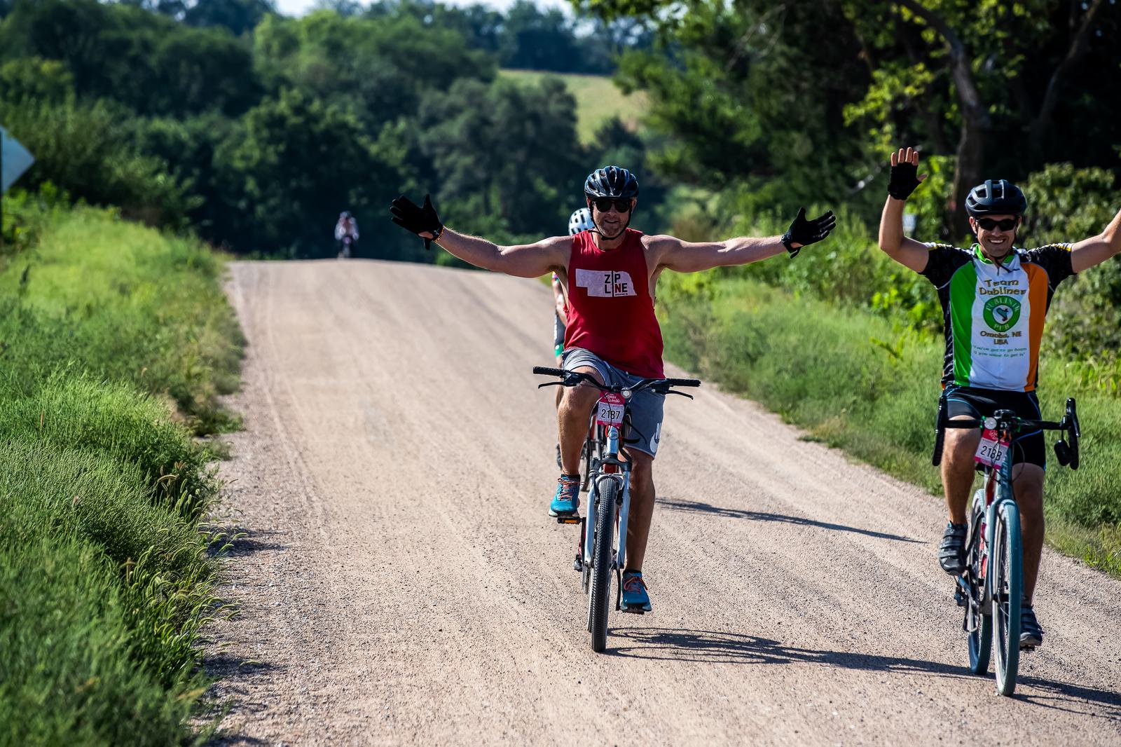 Two cyclists ride on a rural dirt road, smiling with arms raised in celebration. The cyclist on the left wears a red tank top and helmet, while the one on the right wears a green and white jersey. Green foliage and distant trees line the road.