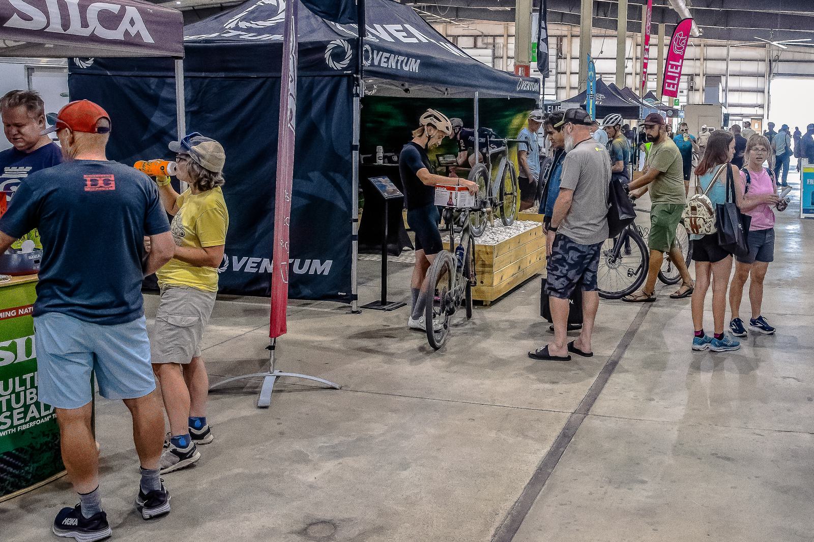 People browse a cycling expo with stalls displaying products. A cyclist in gear walks a bike. Others look at items or chat with vendors. The setting is indoors, with banners and displays set up.