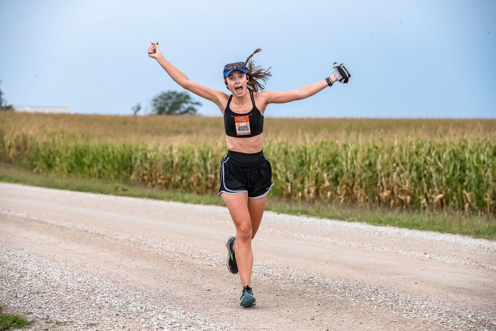 A woman in athletic gear, wearing a visor and numbered bib, runs on a gravel path beside a cornfield. She has her arms raised and a joyful expression, holding a small device in one hand. The sky is overcast.